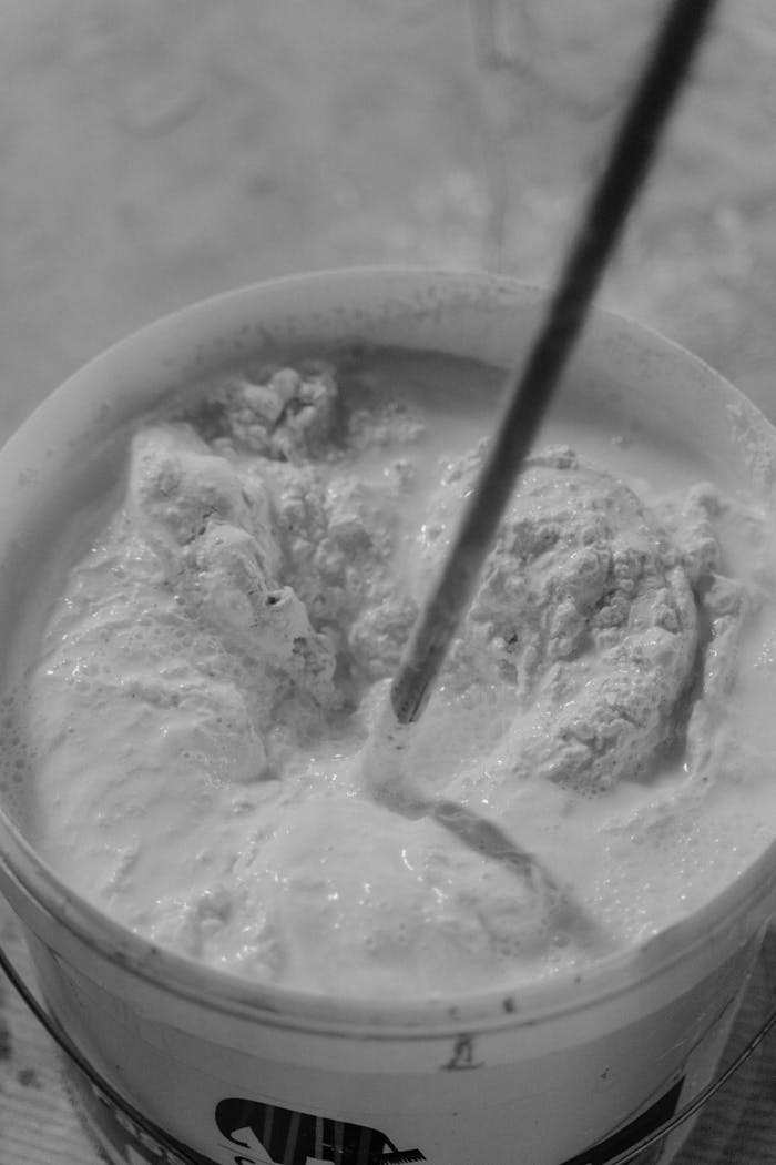 Monochrome image of white paint mixing in a container with a stir stick, showing texture and detail.